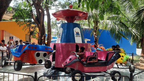 Children enjoying a colorful spinning ride with car-shaped seats at an amusement park, surrounded by trees and excited onlookers.