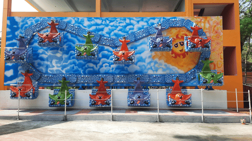 A colourful amusement ride featuring bright animal-shaped seats in red, green, purple, and orange, with a vibrant mural of clouds and a sun wearing sunglasses as the backdrop.