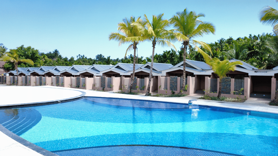The swimming pool area at Symphony Palms Beach Resort And Spa, featuring a curved pool edge, several palm trees, and a row of hut-style villas in the background.