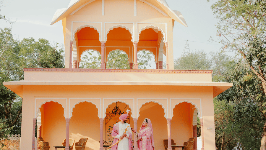 An Indian bride and groom walk together in front of a grand, multi-tiered cream-coloured heritage structure at Umaid Palace.