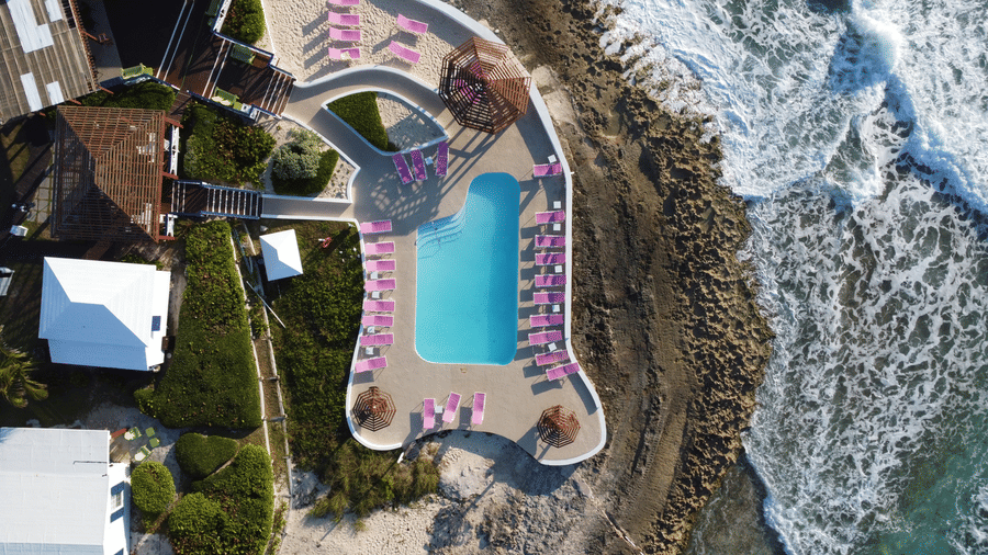 Aerial view of pool in Abaco Inn, with pink lounge chairs, landscaped greenery, and waves crashing against rocky shoreline beside turquoise water