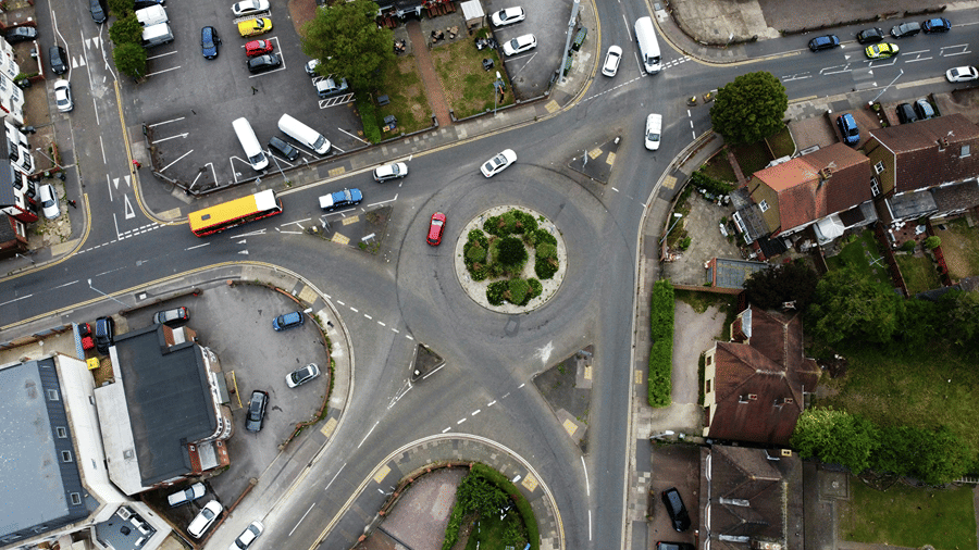 An overhead birds-eye aerial view of a busy urban road roundabout with cars, a yellow bus, and surrounding green trees.