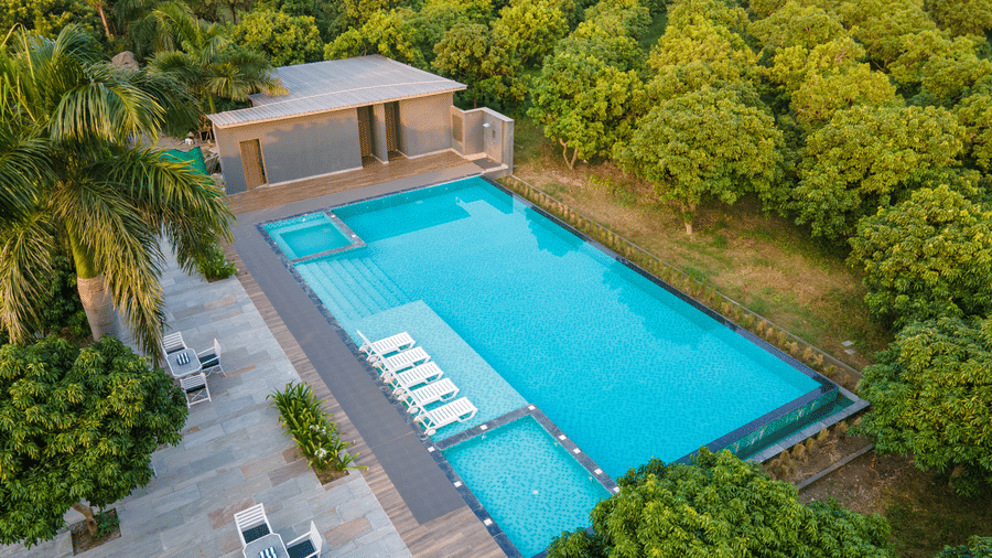An aerial view of a rectangular outdoor swimming pool at Wyndham Garden with a sun deck, lounge chairs, a small structure, and surrounded by trees.