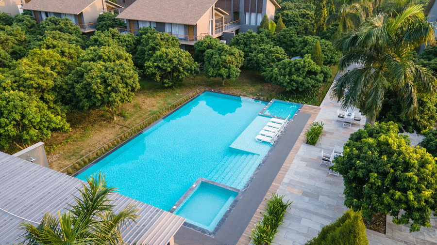 An elevated view of a large outdoor swimming pool at Wyndham Garden with wide entry steps, surrounding deck, and nearby buildings amid dense greenery.