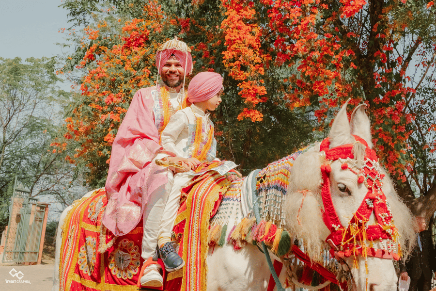 A groom in traditional attire rides a beautifully decorated white horse accompanied by a child during a wedding procession against orange flowers at Umaid Palace.