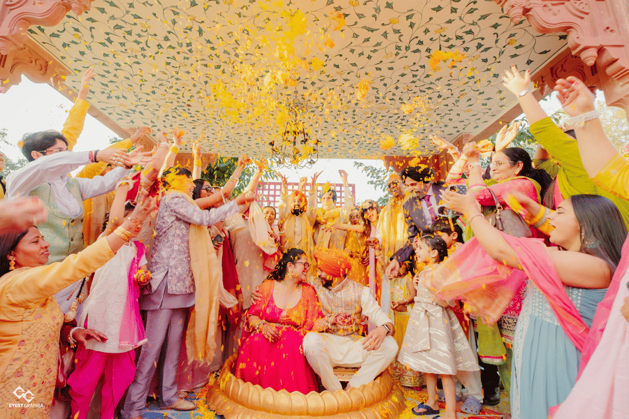 Guests throw yellow flower petals over a couple seated together during a vibrant outdoor Haldi wedding ceremony under a canopy at Umaid Palace.