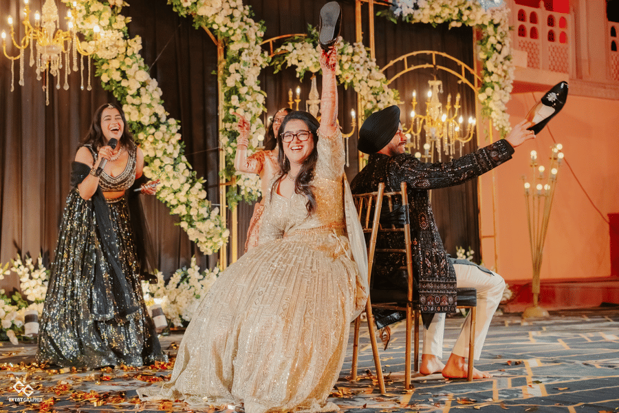 A bride laughs while holding up a shoe during a wedding game on a stage decorated with white flowers and hanging crystals at Umaid Palace.