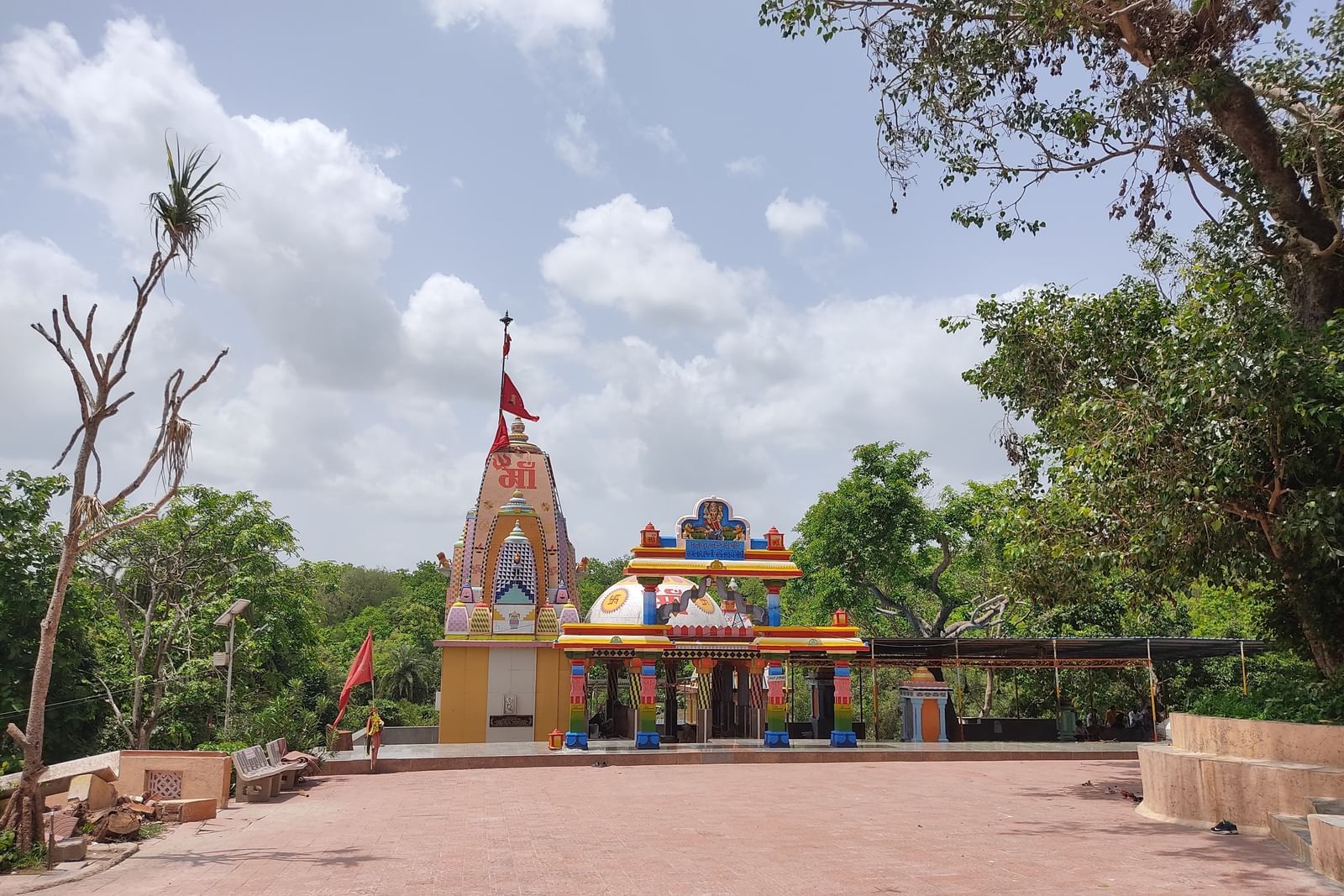 A serene temple painted in bright hues with a tall shikhara and flag, set in an open courtyard surrounded by trees under a partly cloudy sky.