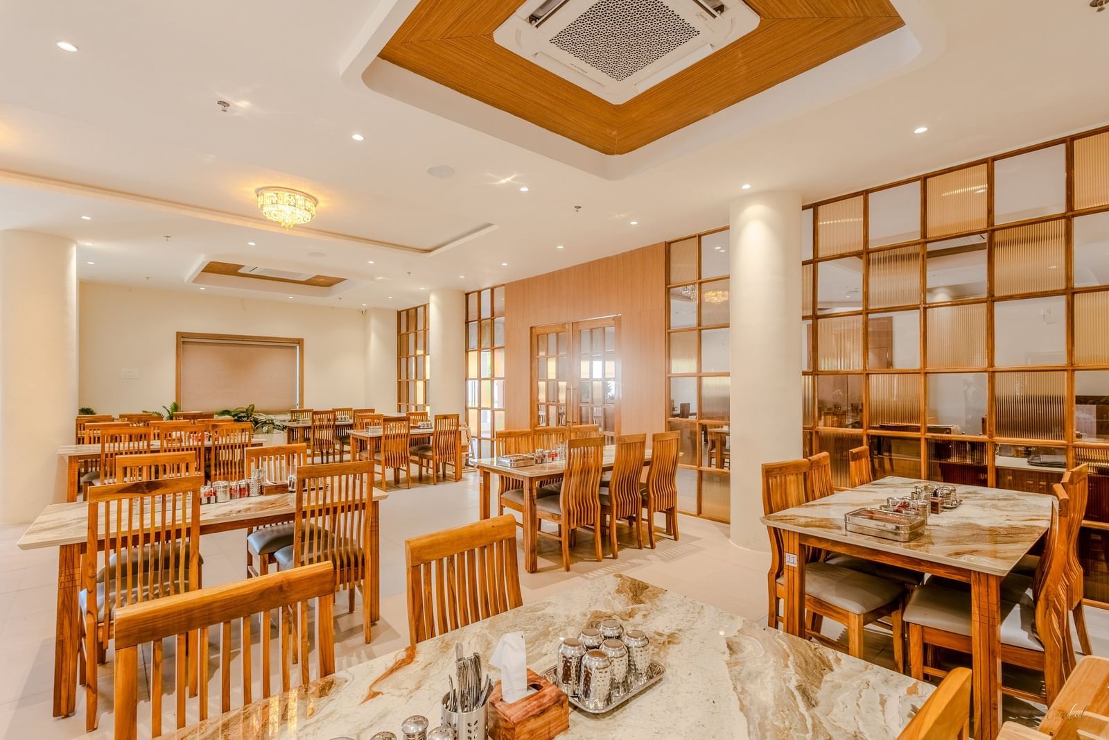 A wide view of the restaurant at Daksh The Grand Gowardhan, Dwarka, showing multiple dining tables and decorative shelving along with central air-conditioning.