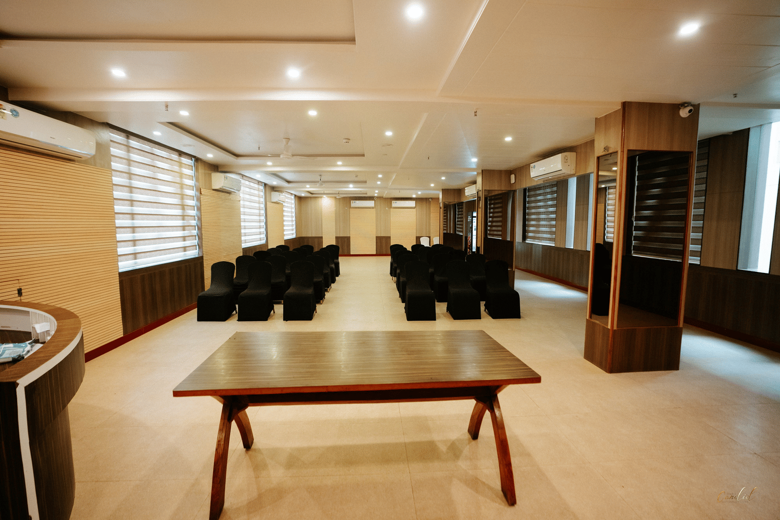 View of the long hall showing a large, dark wood meeting table in the foreground and rows of black chairs further back at Daksh The Madhuvan Suites, Dwarka.