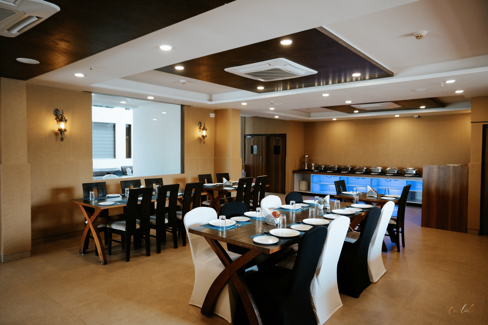 Wide shot of a well-set dining room with dark wood tables, white and black chairs, and beige patterned walls at Daksh The Madhuvan Suites, Dwarka.