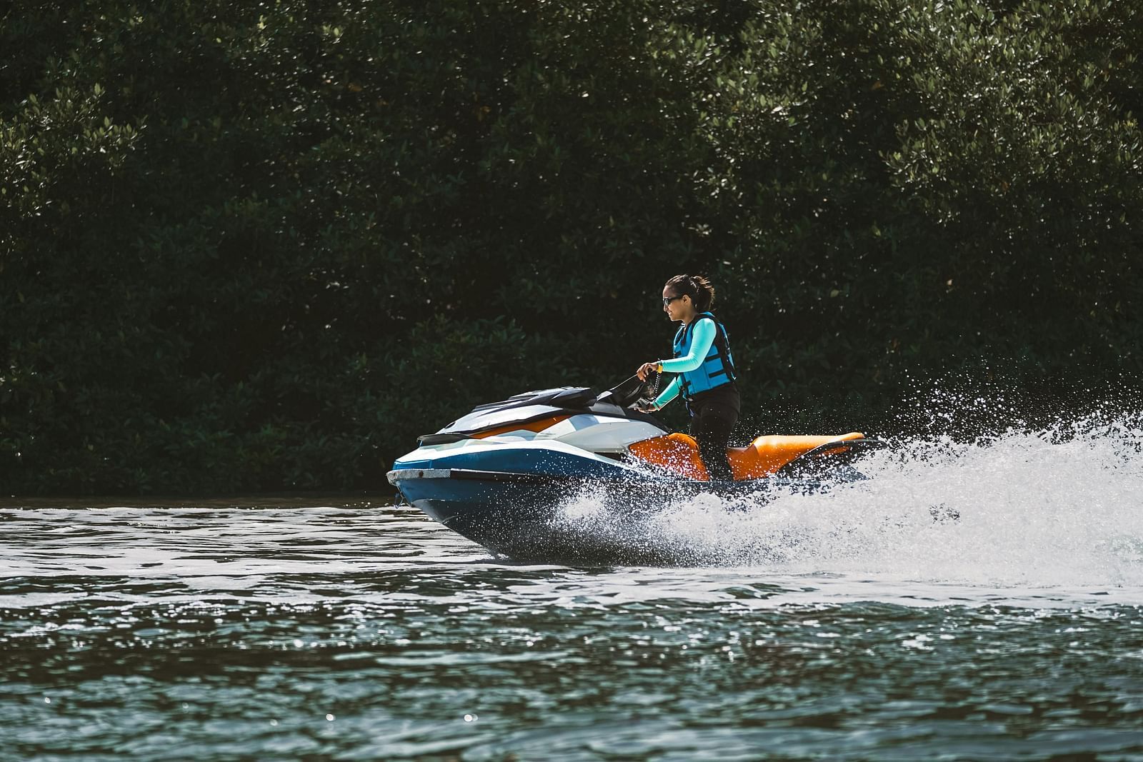 A person in a life jacket is riding a jet ski at speed on a body of water, creating a spray of water, with dense green foliage in the background - Kumarakom
