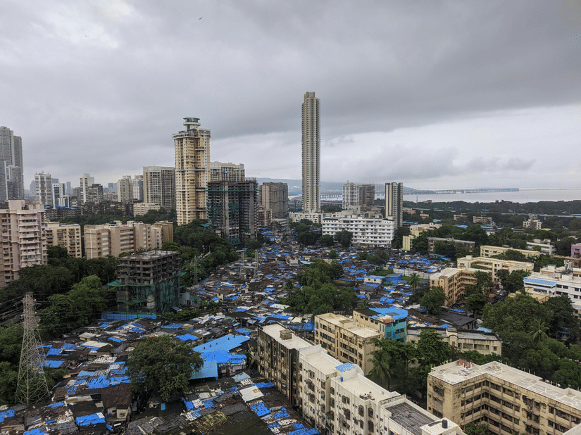 Cityscape with high-rise buildings under cloudy sky, with blue-roofed structures and trees in foreground, and dense urban skyline stretching into distance.