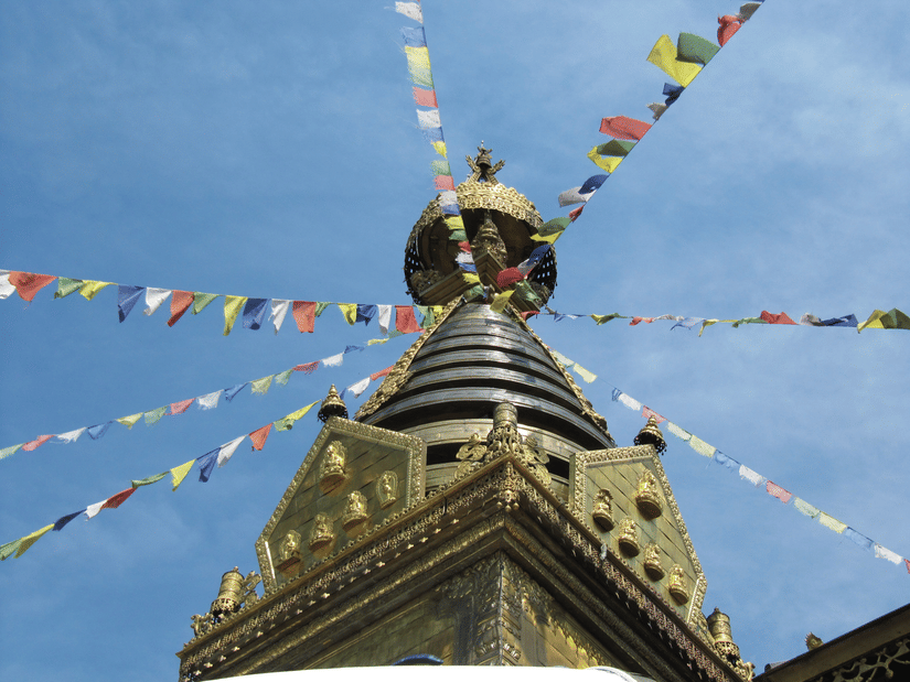 A low-angle view of a golden temple spire with colourful prayer flags strung against a blue sky | HHI Varanasi
