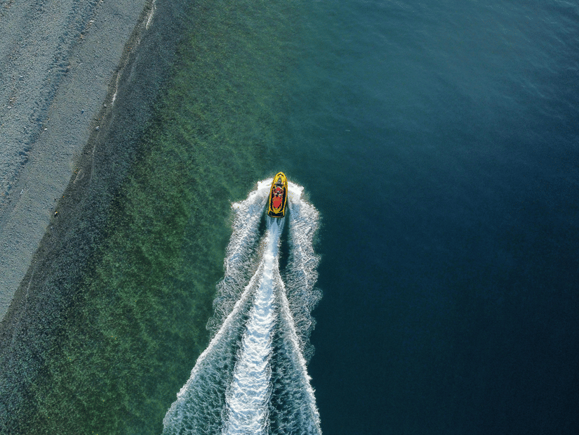 A striking top-down view of a lone speedboat cutting through deep blue ocean waters, leaving a long, foaming white wake behind it.