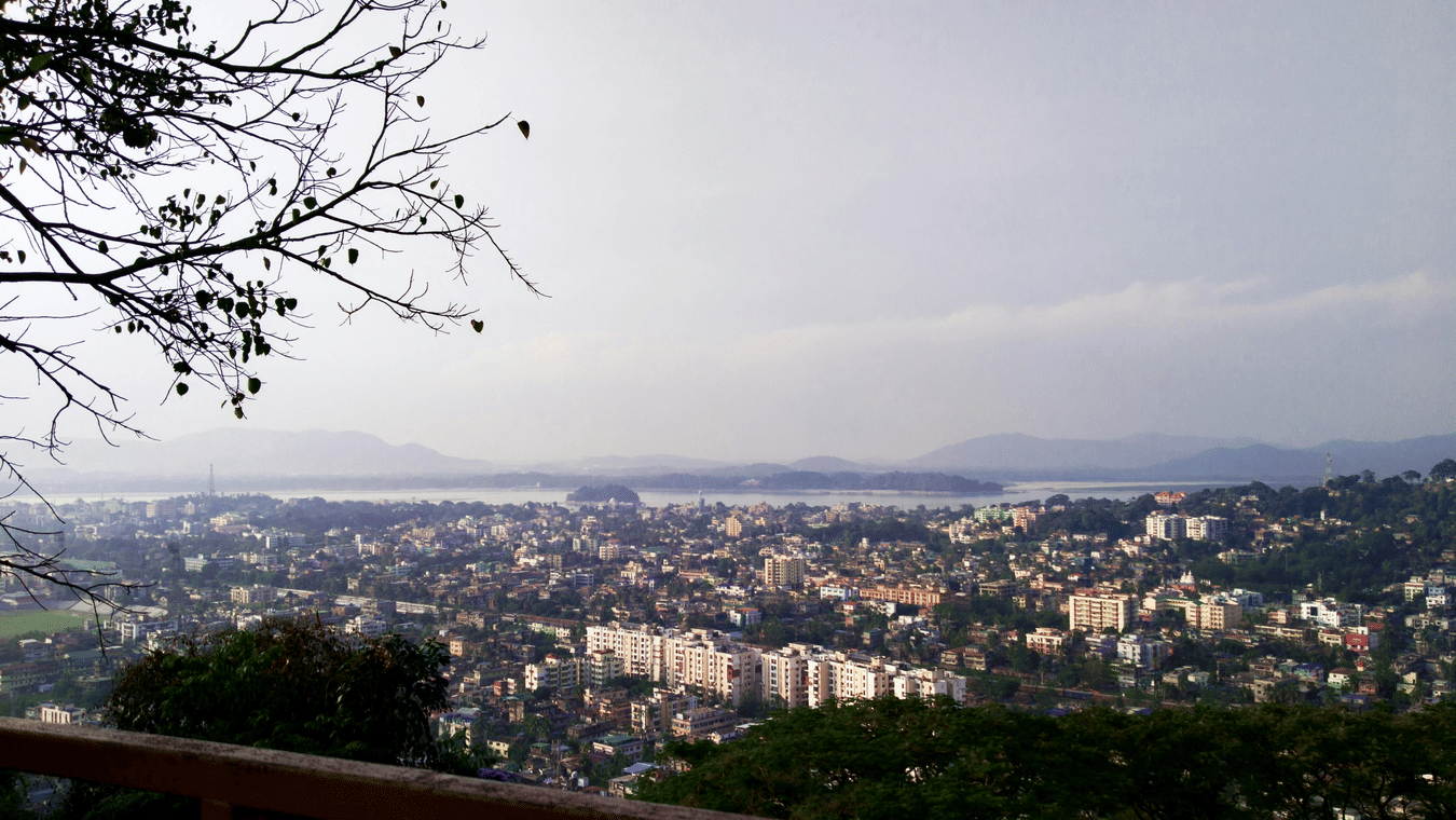 Panoramic view of Guwahati city with dense buildings along the Brahmaputra River, surrounded by distant hills and greenery, seen from an elevated viewpoint under a hazy sky.