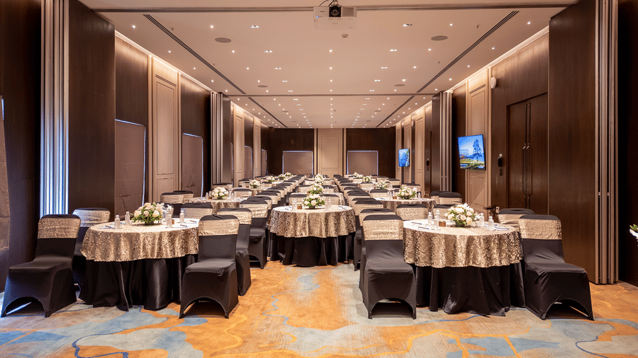 Hotel banquet hall set for an event with rows of round tables draped in striped linens, chairs, and a screen visible at the far end at The Citrine, Bangalore.