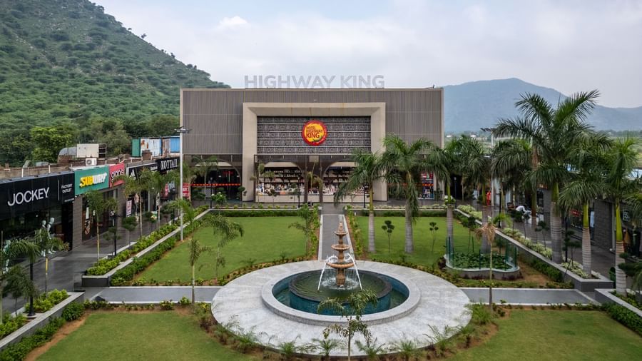 Facade view of Hotel Highway King, Neelka, with a lawn in front, mountains in the background, and shopping complexes on either side.