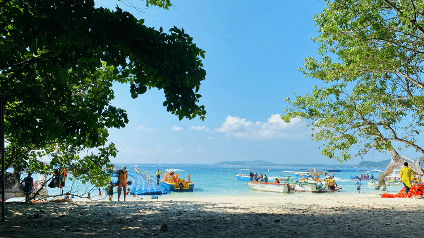 People gather on a sandy beach under leafy green trees as boats wait in the turquoise tropical water.