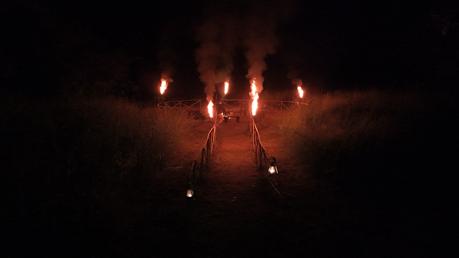 An outdoor night setup with multiple standing fire torches placed in a circular formation on open ground at Infinity Resorts Bandhavgarh.