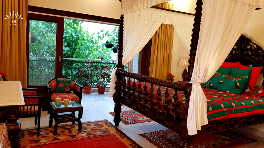 A lavish bedroom showcases an intricately carved four-poster bed with colourful bedding, and a balcony overlooking greenery at Shaheen Bagh Resort and Spa
