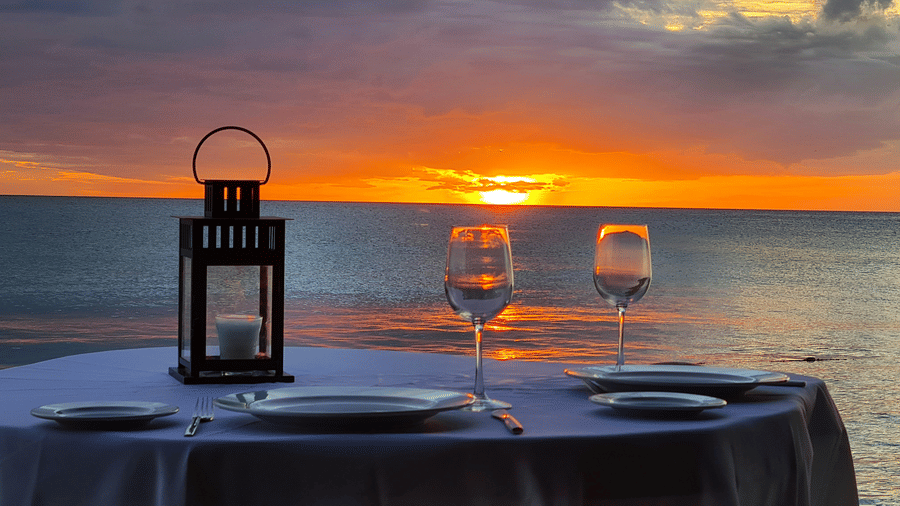 Romantic private dinner table for two with wine glasses and a lantern on the beach during a vibrant orange sunset