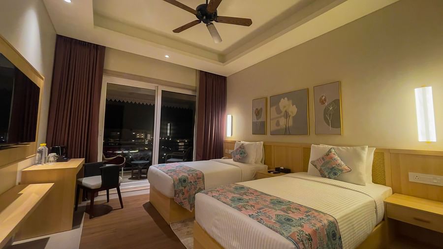 A Room with a balcony having Twin beds with decorated covers and cushions, with a flat screen television, work desk and wall hangings on the room.