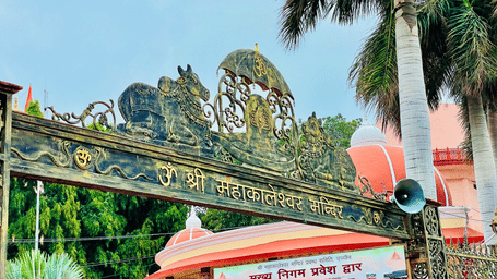 A view of the entrance to Shri Mahakaleshwar Jyotirlinga Temple with a sign board in hindi and Nandis on either side above the signage.