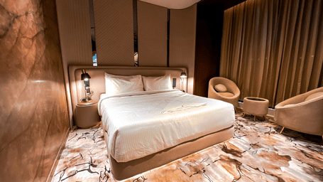 Full view of the double bed with a modern carpet flooring featuring a double bed and a common seating area with 2 arm chairs in the corner at the Honeymoon Suite of F Hotels, Bengaluru.