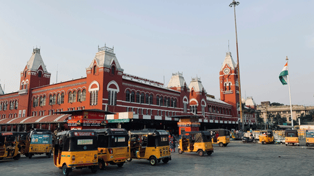 The iconic red Gothic-style facade of Chennai Central Railway Station, featuring its grand clock tower and several yellow auto-rickshaws parked in the foreground.
