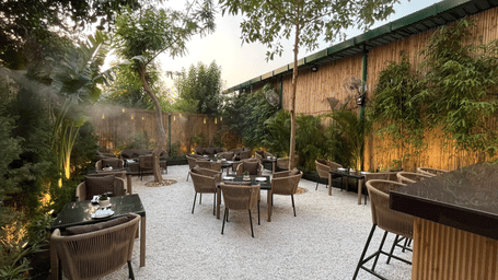 A patio dining space with short round tables, chairs, and a bar table on the side while all of it is surrounded by lush green trees.
