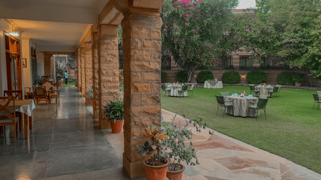 Covered stone verandah with wooden pillars opening to a lush green lawn and outdoor seating at Polo Heritage, Managed by Trance, Jodhpur.