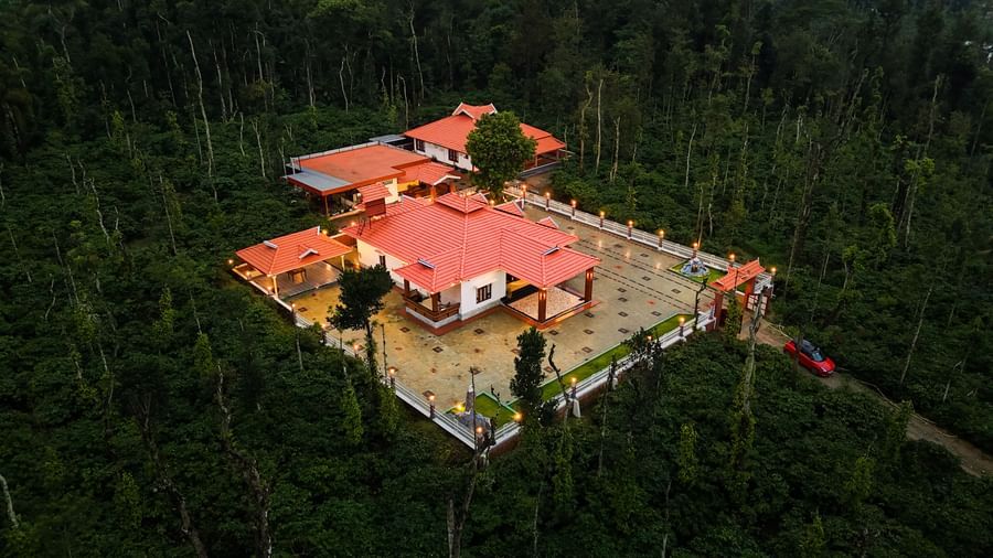 A high-angle aerial shot of the Bhuthanakadu Retreat campus, showcasing the distinct red-tiled roofs of the buildings nestled within the dense, dark green trees.
