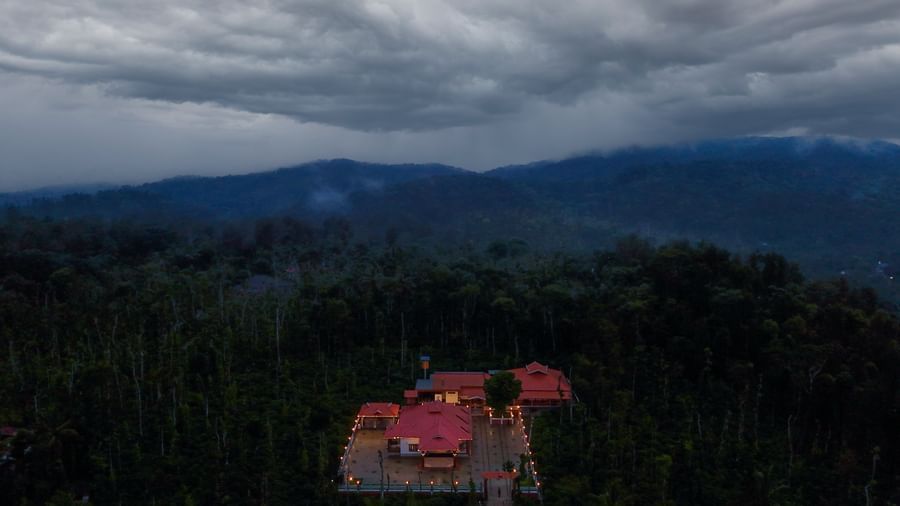 An aerial view of Bhuthanakadu Retreat, with the red-roofed buildings visible at the bottom, surrounded by dark green trees and mist-shrouded hills under a stormy, cloudy sky.