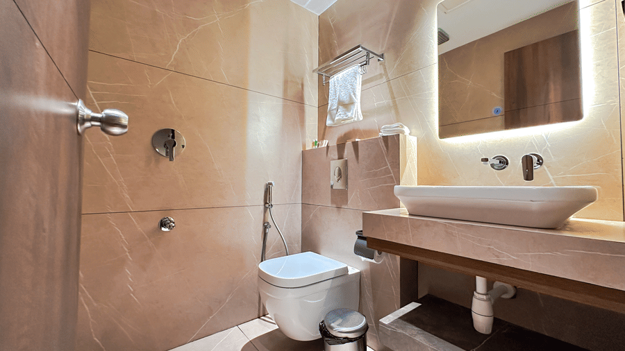 A brightly lit bathroom featuring a showering area, a commode and awash basin with a backlit mirror above it at the Apartment Suite of F Hotels, Bengaluru.