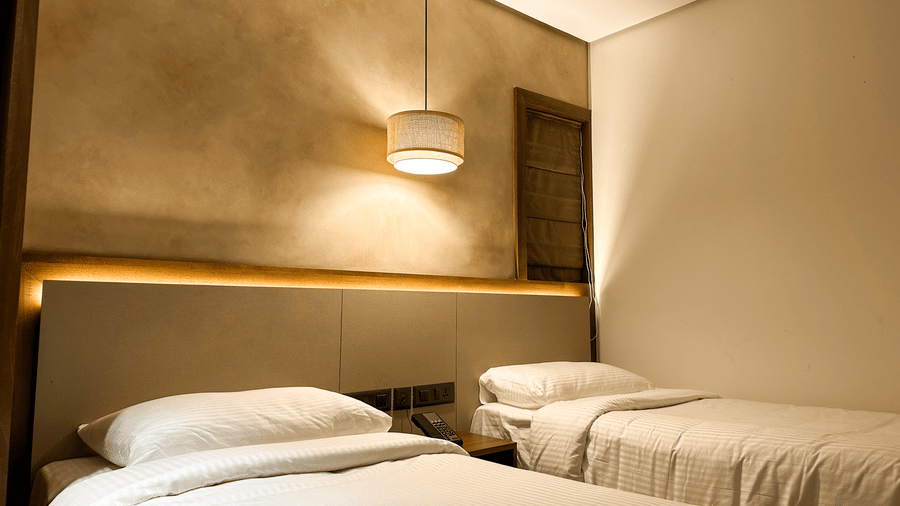 A twin bed set up with a bed side table bewtween the 2 beds, and a modern light fixture hanging from the ceiling in the Deluxe Room at F Hotels, Bengaluru.