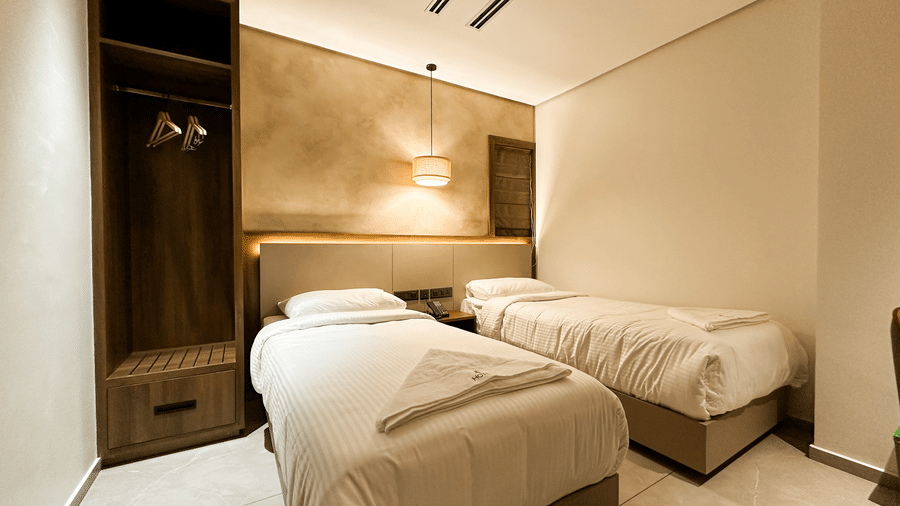 Side view of the Deluxe Room featuring 2 single beds with a backlit headboard along with an open wardrobe in the corner at F Hotels, Bengaluru.