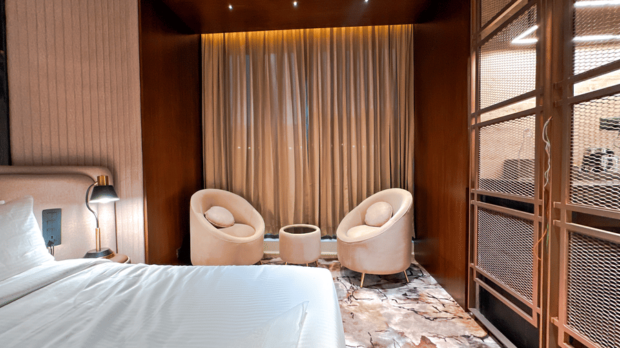 Side view of the bedroom featuring a neatly kept bed beside 2 arm chairs facing each other in the Honeymoon Suite of F Hotels, Bengaluru.