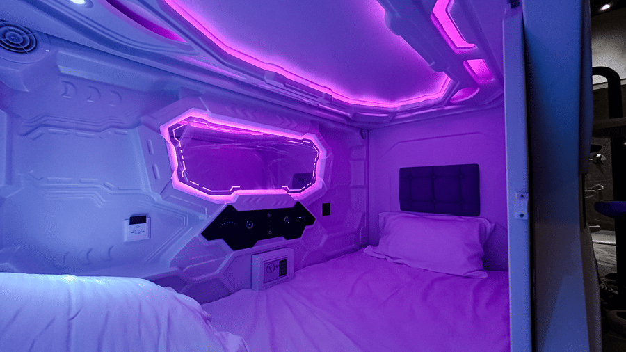 Full view of the interiors of a single private pod with purple lighting featuring a comfortabel bedding and modern amenities at F Hotels, Bengaluru.