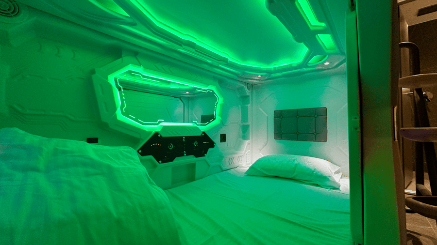 Full view of the pod with green lighting along with a comfortable bedding with a pillow and a comfortor at F Hotels, Bengaluru.