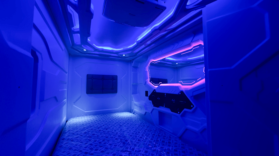 Interior of a pod in F Hotels, Bengaluru, brightly lit blue neon light featuring a small television monitor and wide mirror in one side.