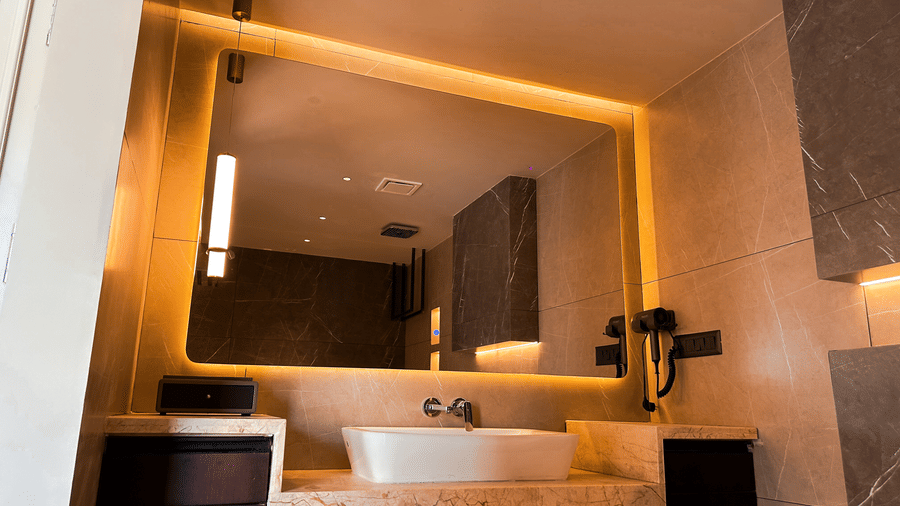 A close-up shot of a huge backlit mirror above the wash basin with a hair dryer mounted on the tiled wall in the bathroom of the Presidential Suite of F Hotels, Bengaluru.