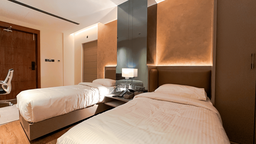 A wide-angle shot of the Superior Room at F Hotels, Bengaluru, featuring nealty kept twin beds with a pillar covered with mirror pannels in the middle of the 2 beds.