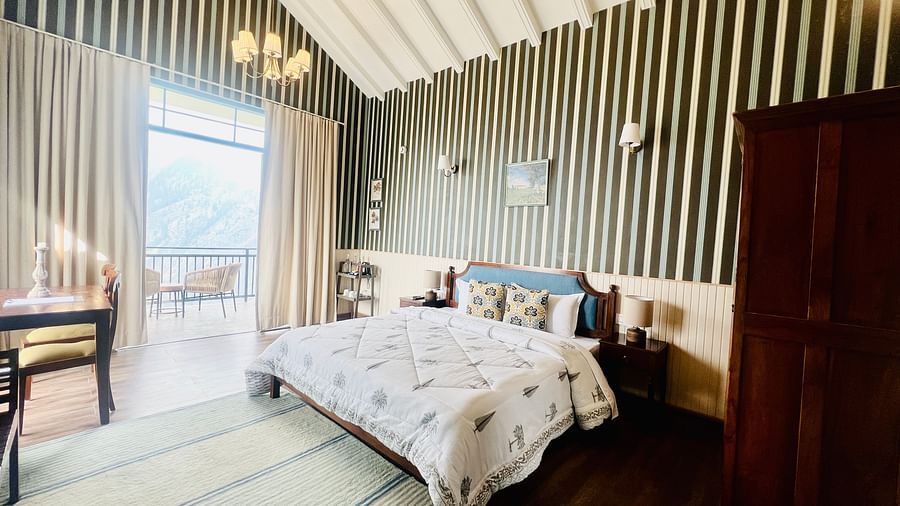 Spacious hotel room featuring a wooden bed with white and gray bedding, striped walls, and a panoramic window with mountain views | Faraway Cottages, Auli