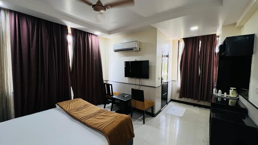  A comfortable hotel bedroom with a large bed, a gold-colored bed runner, a television, and dark curtains | Deluxe Room | Hotel Aadhar
