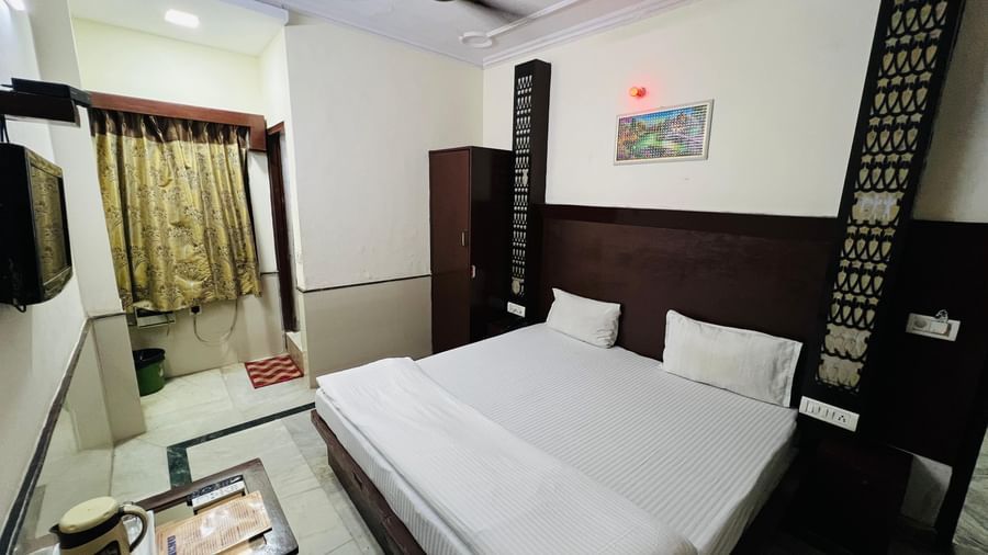  A well-lit room with a comfortable bed, a small table, a TV, and patterned curtains, offering a cozy and functional space. | Deluxe Room | Hotel City Palace DX