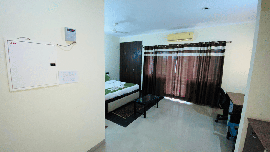 A view of a bed partially visible in a guest room featuring an AC, wardrobe, a table and brown curtains drawn across the window at Livbox Rudrapur.