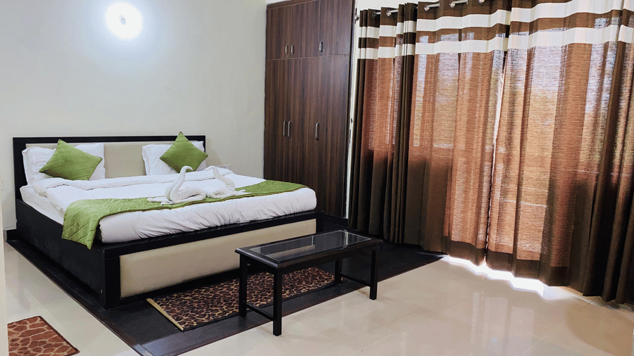 A bedroom with a green-accented bed, long brown curtains, a rug and a glass-topped table at Livbox Rudrapur.