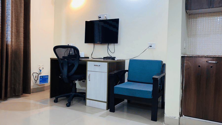 A work corner with a black office chair, mounted TV, white drawer unit, and a blue cushioned chair at Livbox Rudrapur.