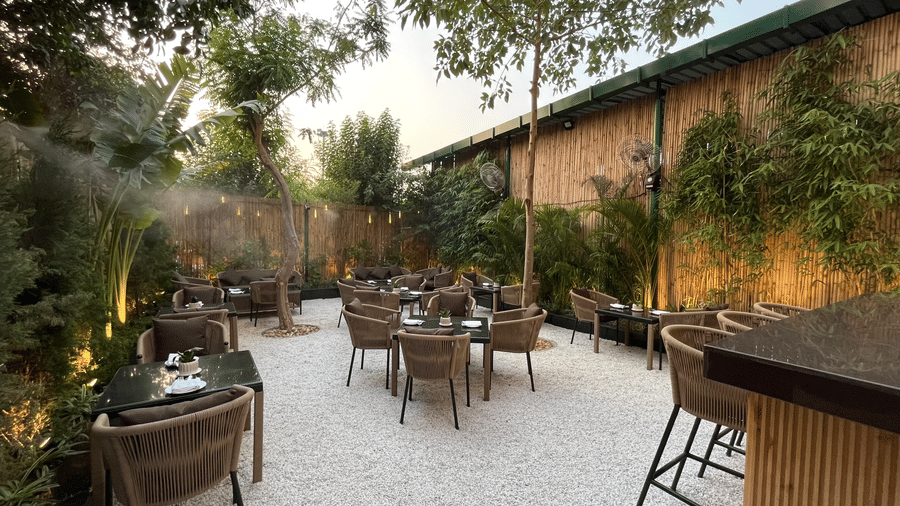 A patio dining space with short round tables, chairs, and a bar table on the side while all of it is surrounded by lush green trees.